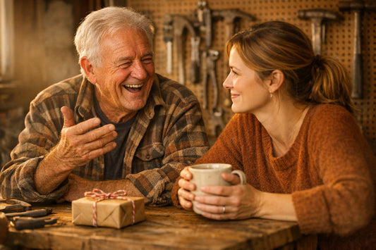 A father laughing as he tells his adult daughter a story at his garage workbench, a small wrapped gift beside them